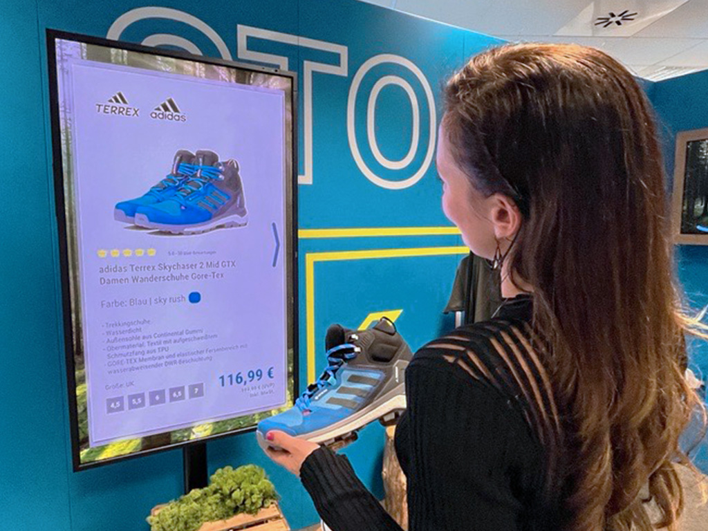 Kundin im Sportgeschäft hält einen Wanderschuh und betrachtet auf einem Bildschirm die Produktdetails und den Preis.