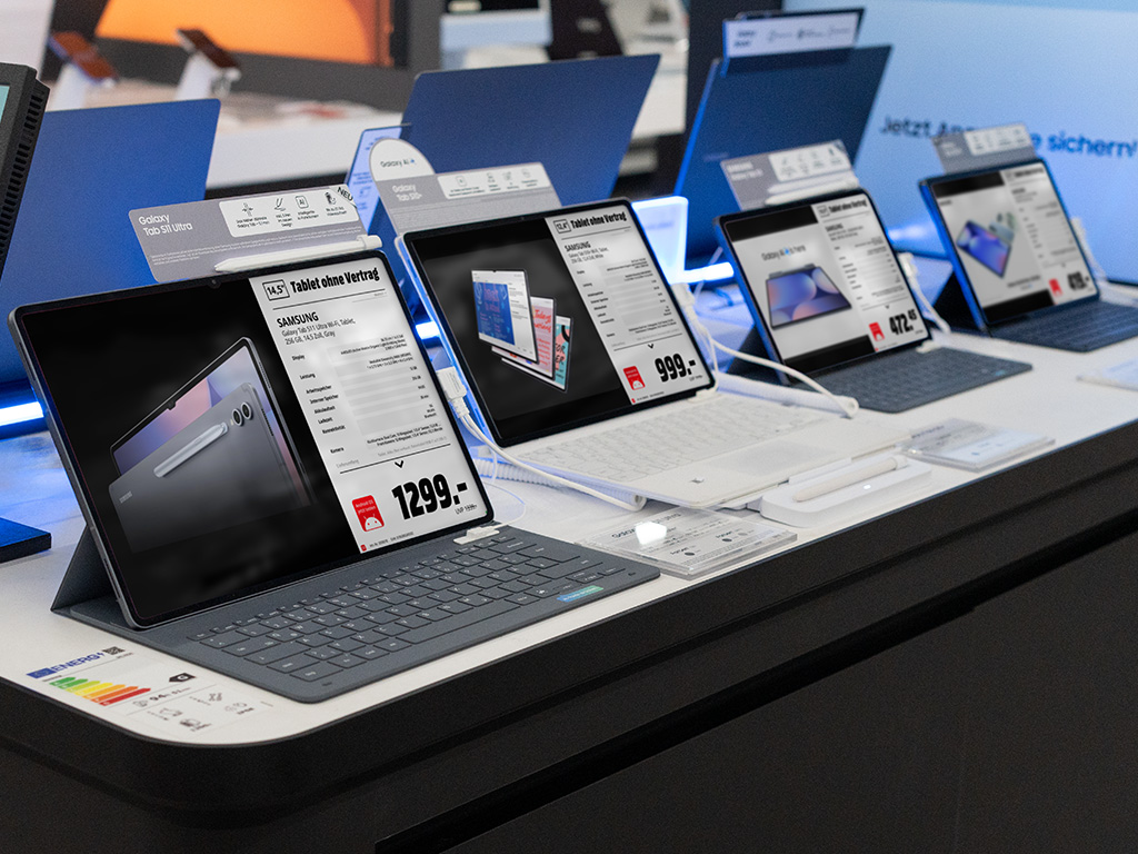 Ausstellungsfläche im Elektronikmarkt mit mehreren Tablets/Laptops; auf den Bildschirmen sind digitale Produktdatenblätter und Preise zu sehen.